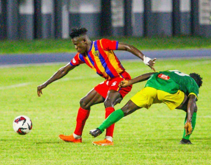 Soccer players contesting the ball on a green field; one in a red-yellow striped jersey and red shorts, the other in green and yellow.