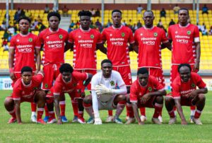 Soccer team in red uniforms posing for a group photo on a green field with yellow stadium seats in the background.
