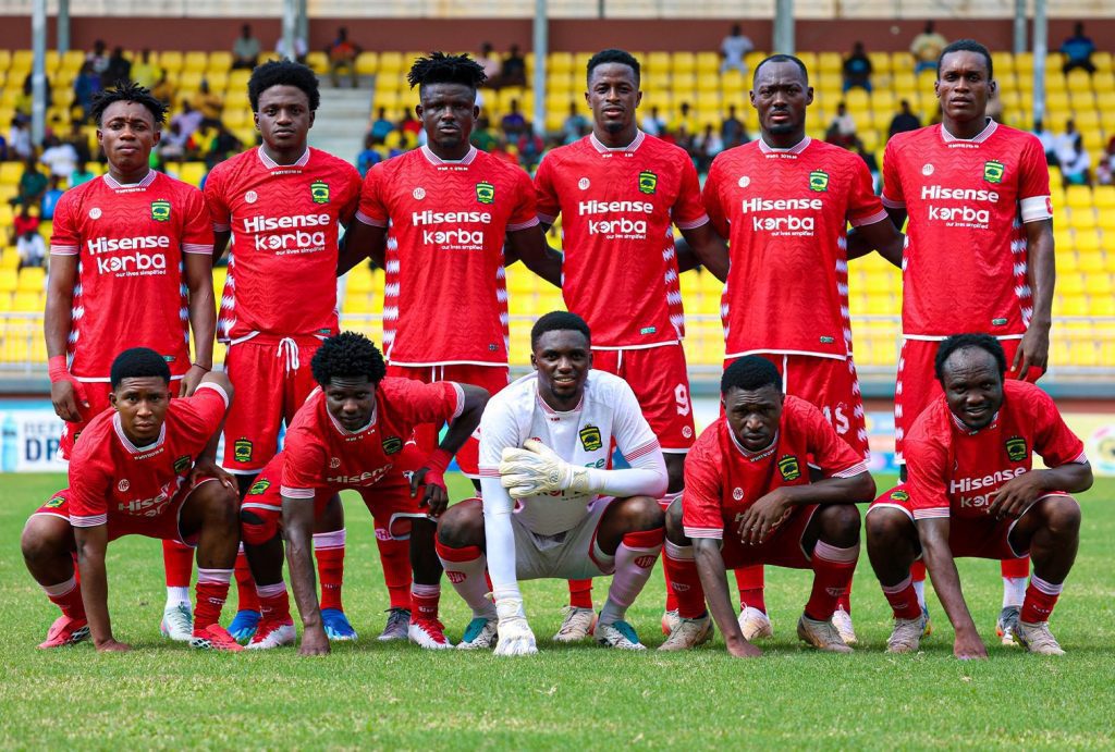 Soccer team in red uniforms posing for a group photo on a green field with yellow stadium seats in the background.
