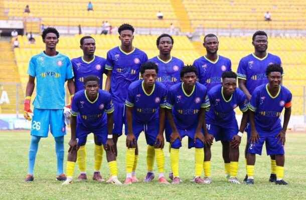 Soccer team posing for a group photo on a grassy field, players in blue and yellow uniforms with a goalkeeper in light blue stand at the back in a stadium with yellow seats.
