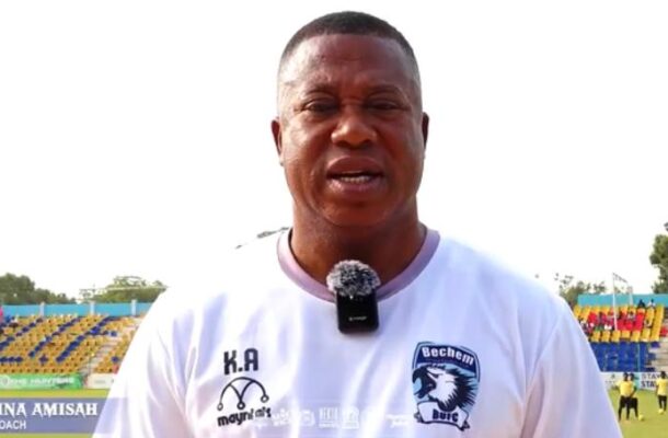 Head coach in a white Bechem United shirt speaks into a lavalier mic on a football pitch with colorful stadium seating in the background