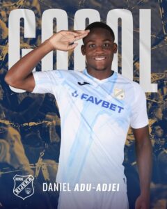 Portrait of soccer player Daniel Adu-Adjai saluting, wearing a light blue and white jersey with Favbet sponsor; team crest visible, cheering crowd backdrop and a large 'GOAL' graphic behind him.