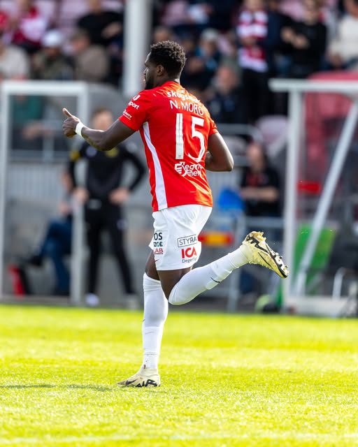 Soccer player wearing a red jersey with the number 15, running on a sunny grass field during a match, with spectators blurred in the background and white socks and cleats visible.