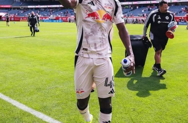 Soccer player in a white Red Bull kit waves to the crowd on the pitch, holding a water bottle.