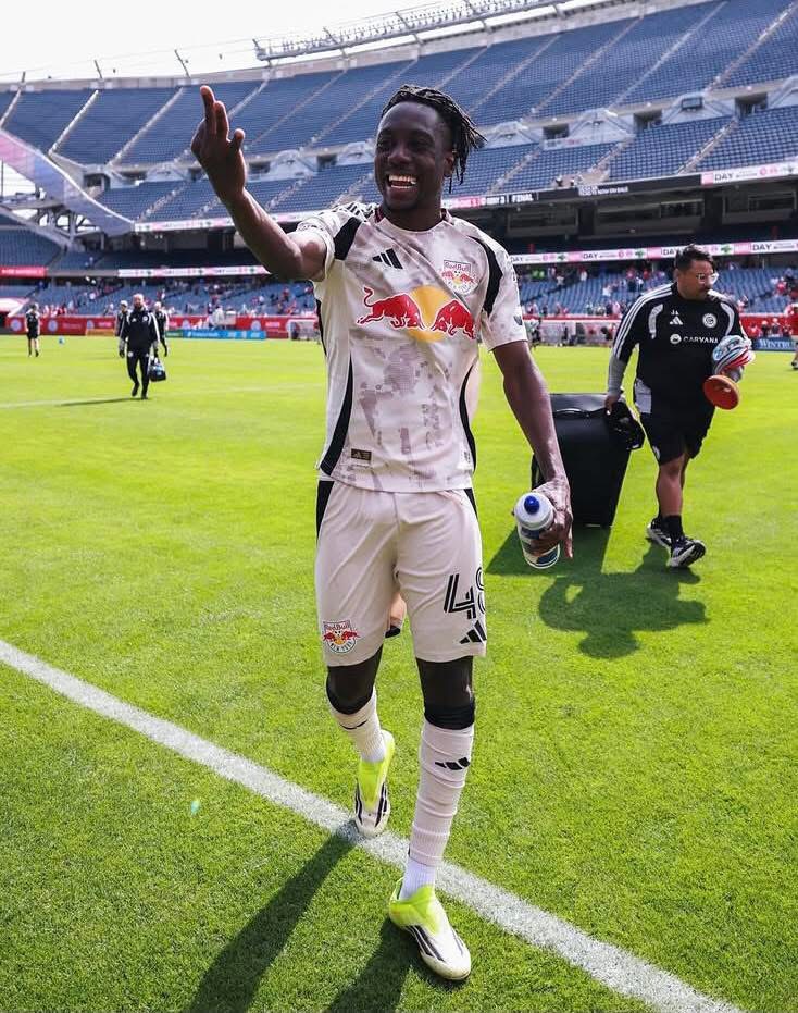 Soccer player in a white Red Bull kit waves to the crowd on the pitch, holding a water bottle.