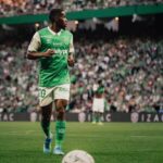 Soccer player in a green and white kit on a packed stadium field, ball at his feet before a kickoff or pass.