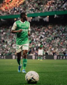 Soccer player in a green and white kit on a packed stadium field, ball at his feet before a kickoff or pass.