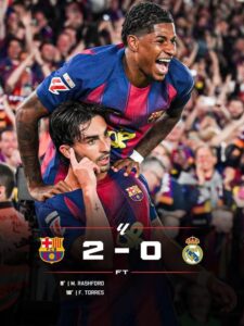 Two FC Barcelona players celebrate a 2–0 victory over Real Madrid as fans cheer in the background, scoreboard showing 2–0.