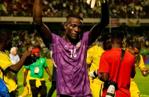Soccer goalkeeper in purple kit (#16) raises arms in victory on a crowded stadium field, gloves in the air.