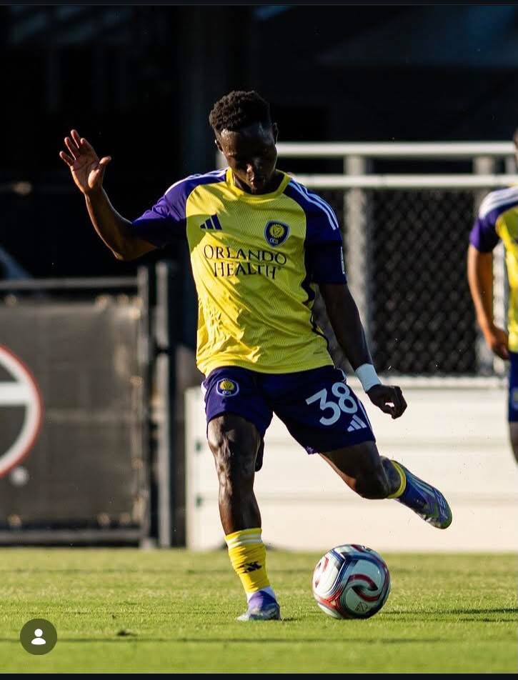 Soccer player in yellow and purple jersey (Orlando Health) about to kick a white ball on a grassy field; number 38 on shorts, raised left hand.