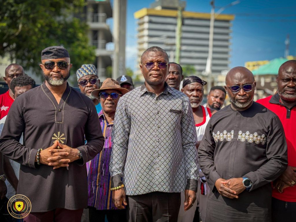 Group of men standing together outdoors for a photo, with buildings in the background.