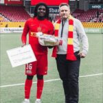 Football player in red kit holding a 'Man of the Match' certificate and a trophy, standing with a man in a red-and-yellow scarf on a stadium pitch.
