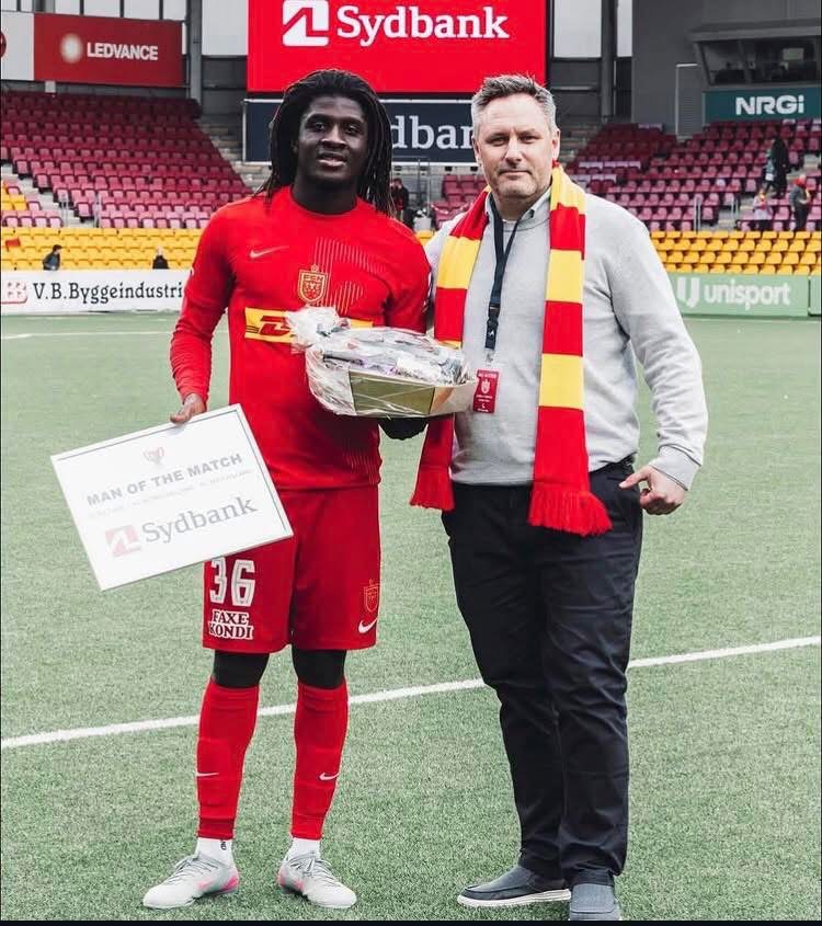 Football player in red kit holding a 'Man of the Match' certificate and a trophy, standing with a man in a red-and-yellow scarf on a stadium pitch.