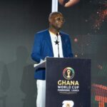 Man in a blue blazer speaks at a podium during the Ghana World Cup Fundraising Launch event with a crest on the podium.