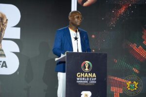 Man in a blue blazer speaks at a podium during the Ghana World Cup Fundraising Launch event with a crest on the podium.