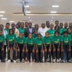 Large group photo of around 50 people wearing matching green t-shirts, posing together indoors in a spacious terminal or lobby area.