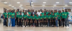Large group photo of around 50 people wearing matching green t-shirts, posing together indoors in a spacious terminal or lobby area.