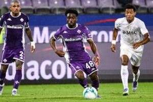 Three soccer players chase the ball on a green pitch; two wear purple Fiorentina jerseys, one in white, during a match.