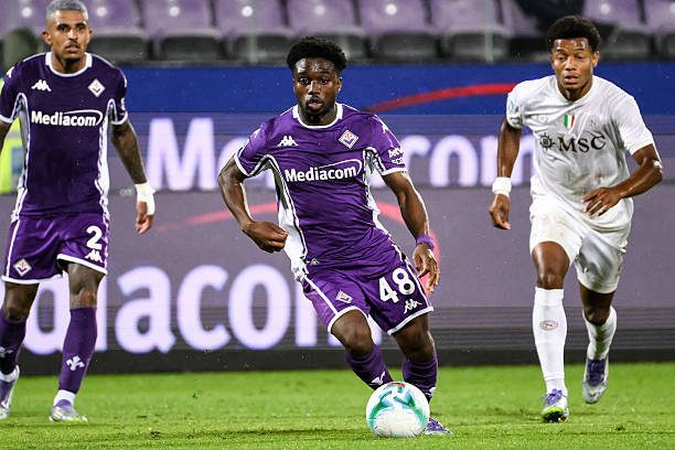 Three soccer players chase the ball on a green pitch; two wear purple Fiorentina jerseys, one in white, during a match.