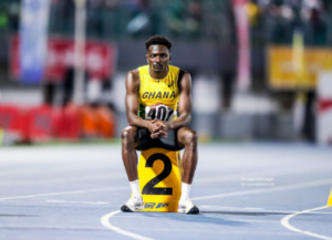 Ghanaian sprinter in a yellow uniform crouched at the starting line on a track, wearing a large number 2 bib, ready to start the race.