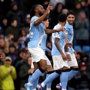 Soccer players in light blue jerseys celebrate a goal on the pitch with fans in the stands behind them.