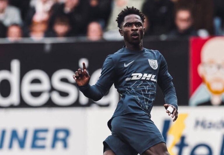 Soccer player in a navy kit with DHL logo runs on the pitch, with a blurred crowd in the background.