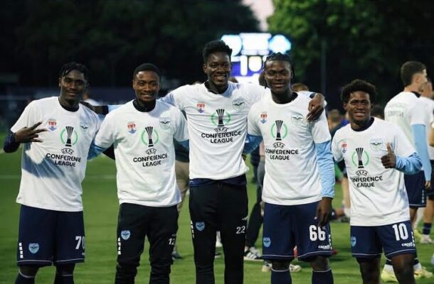 Five male soccer players in white Conference League jerseys pose on a field, arms around each other and smiling.