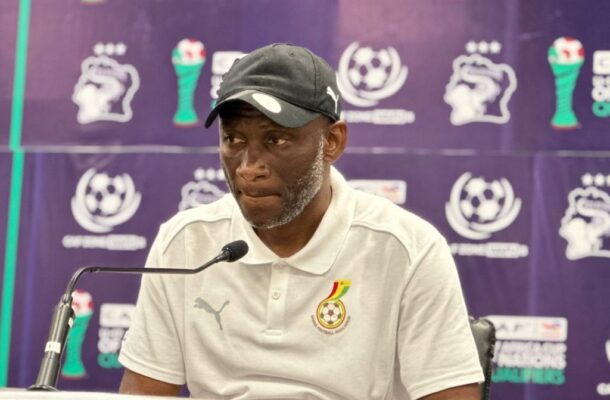 Man in a white polo shirt and black cap speaks into a microphone at a football press conference, with a purple branded backdrop behind him.