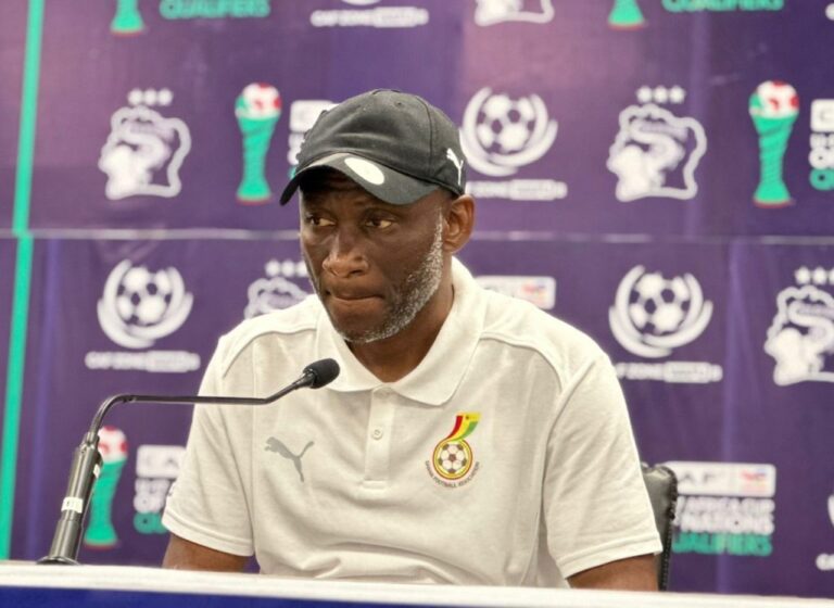 Man in a white polo shirt and black cap speaks into a microphone at a football press conference, with a purple branded backdrop behind him.
