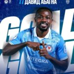 Football player in a light blue jersey smiling and making a celebratory hand gesture for a club poster or promo image.