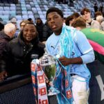 Young soccer player in a light blue jersey holds a silver trophy, with a scarf draped over his shoulders, as fans cheer in the stands behind him.