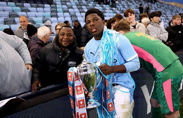 Young soccer player in a light blue jersey holds a silver trophy, with a scarf draped over his shoulders, as fans cheer in the stands behind him.