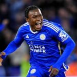 Leicester City footballer in a blue kit celebrates a goal on the pitch, mouth open in celebration, with a blurred crowd in the background.
