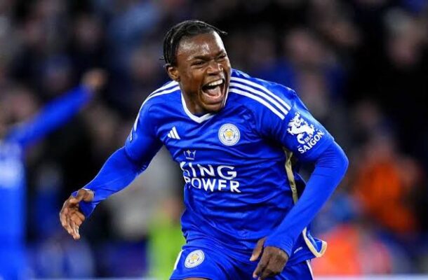 Leicester City footballer in a blue kit celebrates a goal on the pitch, mouth open in celebration, with a blurred crowd in the background.