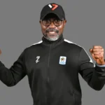 Smiling man in a black athletic jacket and cap, fists raised in victory against a gray background.