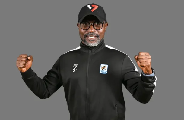 Smiling man in a black athletic jacket and cap, fists raised in victory against a gray background.