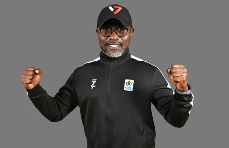 Smiling man in a black athletic jacket and cap, fists raised in victory against a gray background.