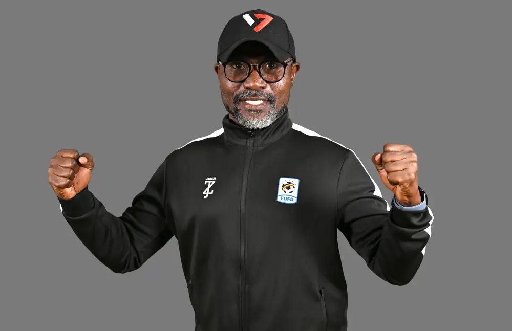 Smiling man in a black athletic jacket and cap, fists raised in victory against a gray background.