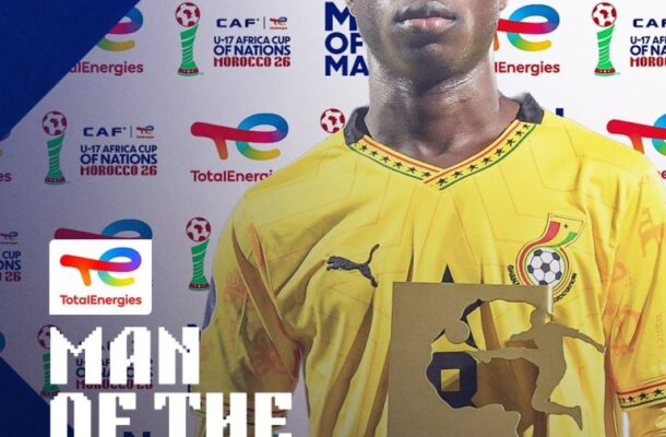 Eric Gyamfi in a yellow jersey holding the Man of the Match trophy at a sponsor backdrop.