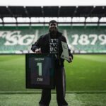 Man on a soccer field holding a framed jersey with the number 1 and the name ZIGI, beside a plaque reading Key Player.