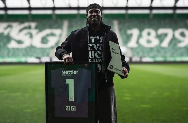 Man on a soccer field holding a framed jersey with the number 1 and the name ZIGI, beside a plaque reading Key Player.