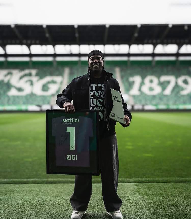 Man on a soccer field holding a framed jersey with the number 1 and the name ZIGI, beside a plaque reading Key Player.
