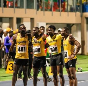 Four Ghanaian male runners in yellow, black shorts, with their arms around each other after a race, posing on a track with bib numbers 401 and 1400 visible.