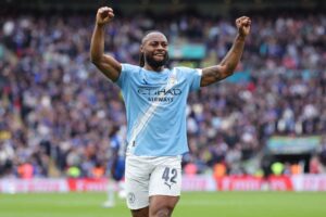 Manchester City player (number 42) celebrates a goal with fists raised on the field, crowd blurred in the background.