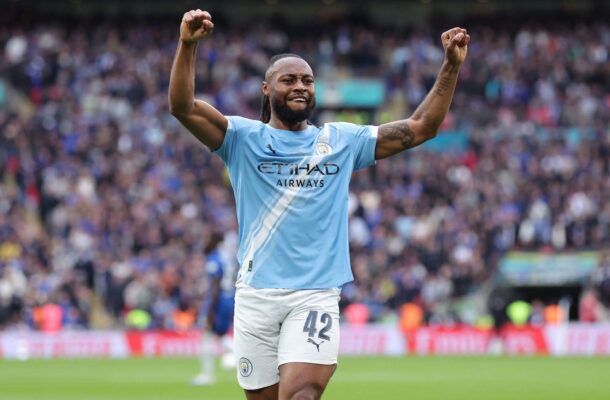 City footballer celebrates with arms raised on the pitch as a crowded stadium watches in the background, wearing a light blue kit and number 42