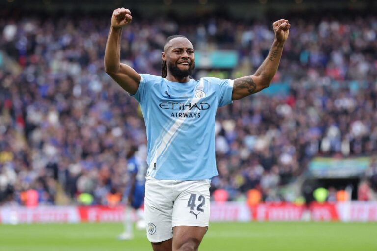 City footballer celebrates with arms raised on the pitch as a crowded stadium watches in the background, wearing a light blue kit and number 42