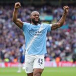 Soccer player in Manchester City kit celebrating with arms raised amid a cheering crowd in a stadium.