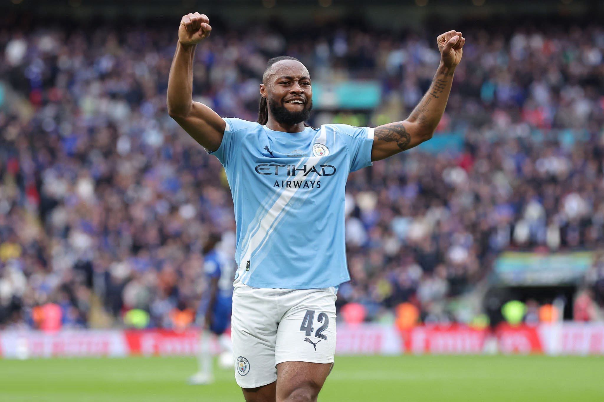 Soccer player in Manchester City kit celebrating with arms raised amid a cheering crowd in a stadium.