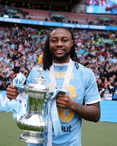 Soccer player in a light-blue jersey smiles while holding a large silver trophy with light-blue ribbons, in a stadium full of fans behind him.