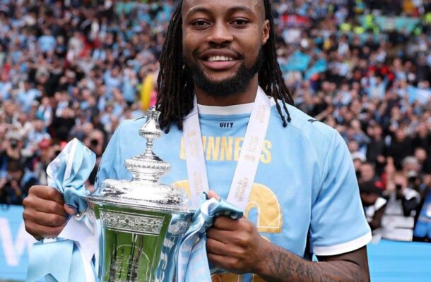 Soccer player in a light-blue jersey smiles while holding a large silver trophy with light-blue ribbons, in a stadium full of fans behind him.